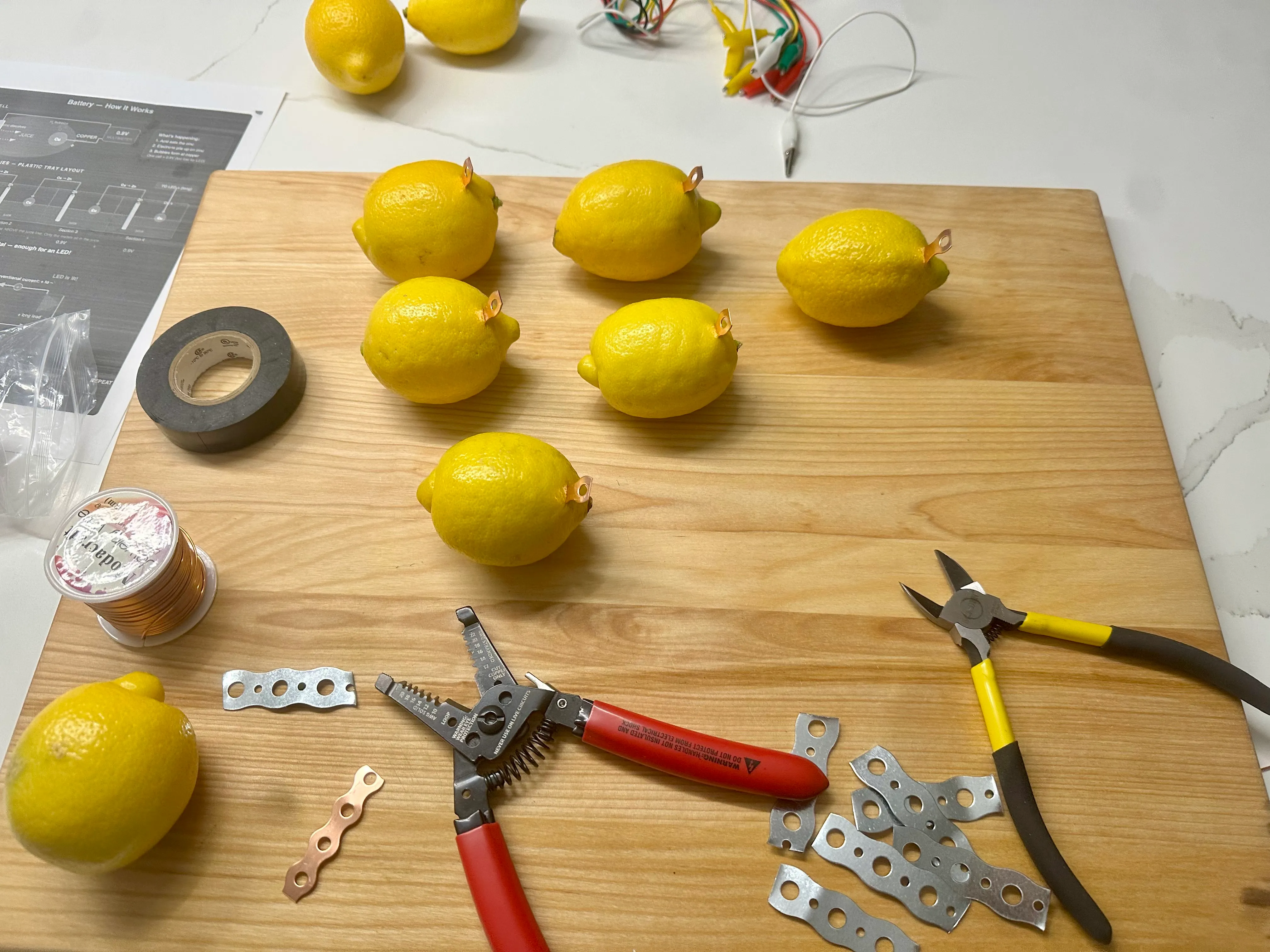 Lemons with copper electrodes on a cutting board, surrounded by galvanized brackets, wire strippers, electrical tape, and a spool of copper wire