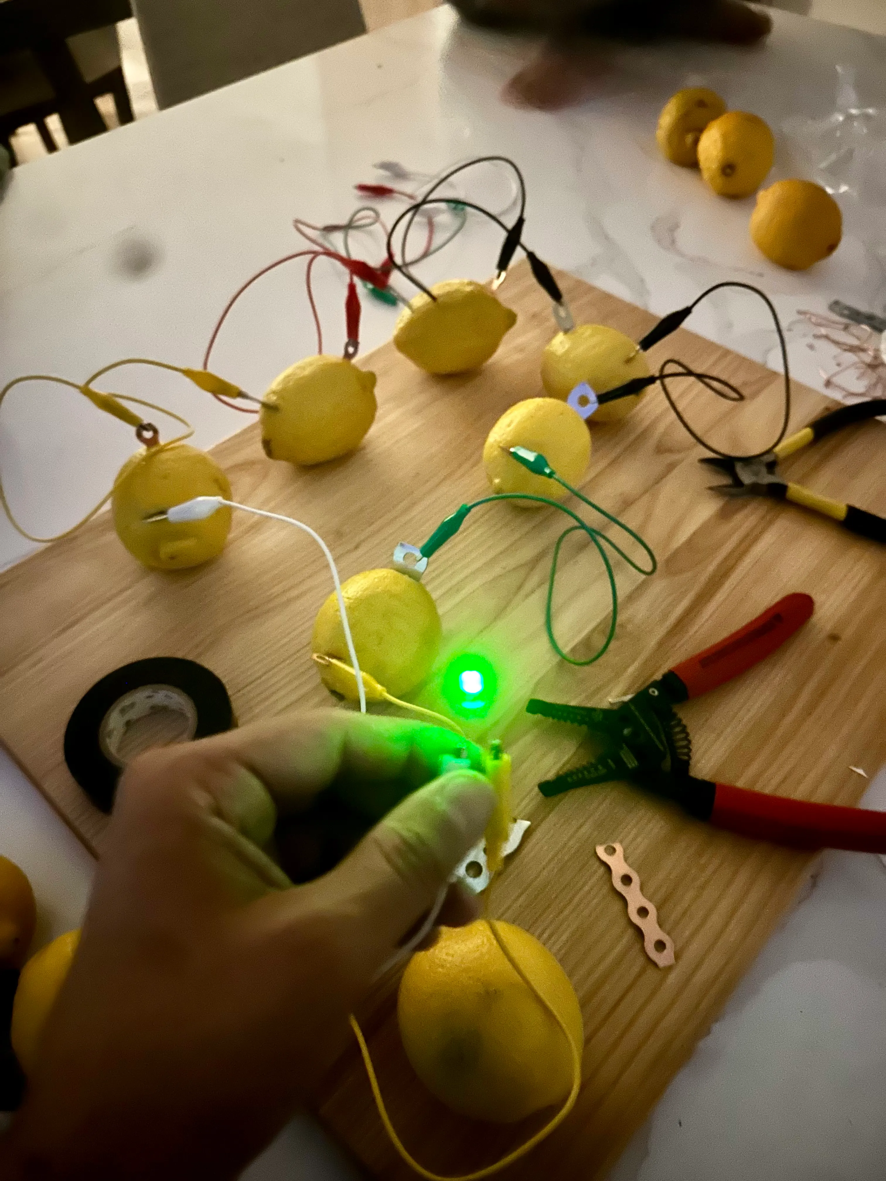 Chain of lemons connected with alligator clip leads on a cutting board, a green LED glowing in hand
